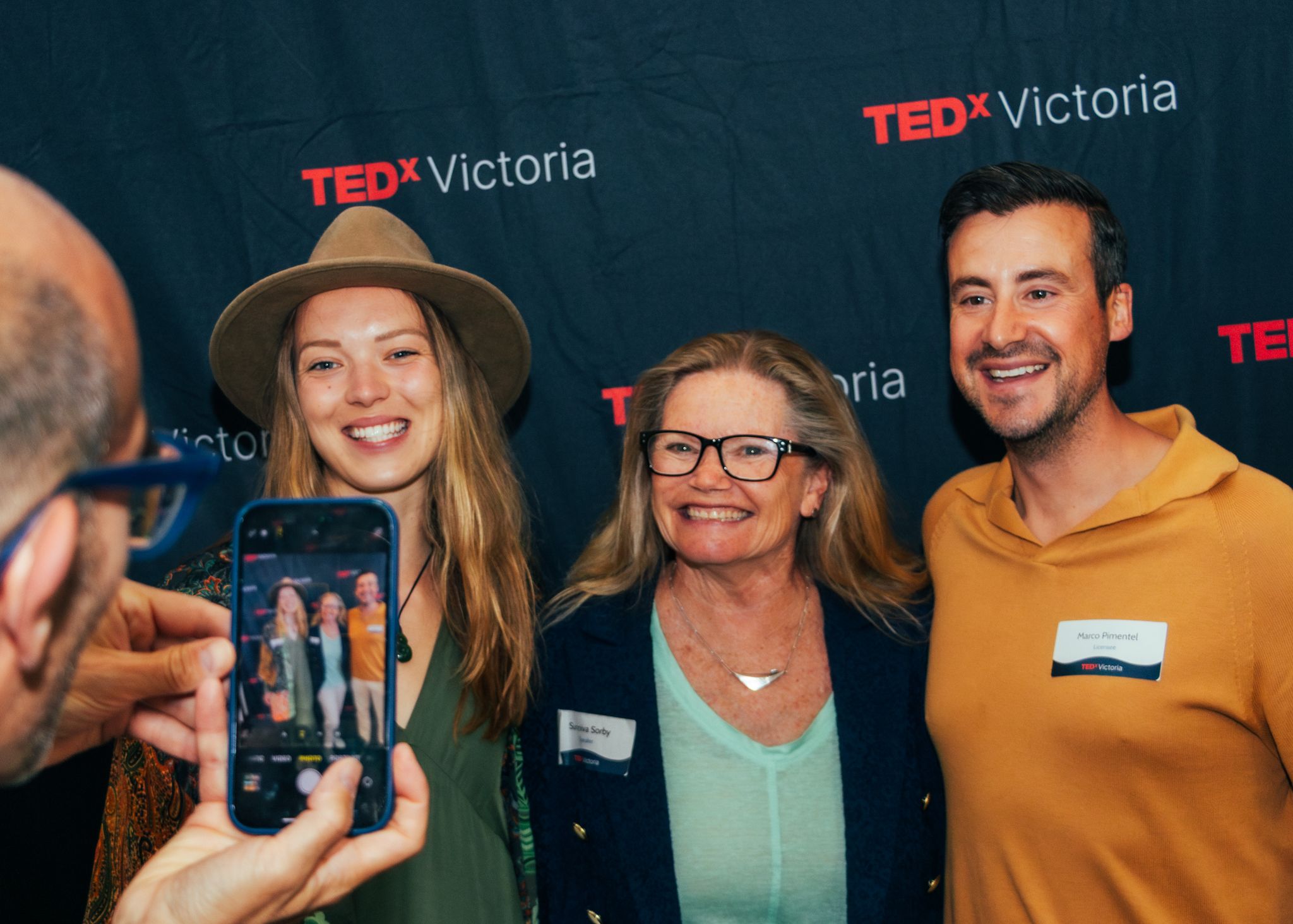 Three people pose for a photo in front of a TEDx Victoria backdrop at a networking event.