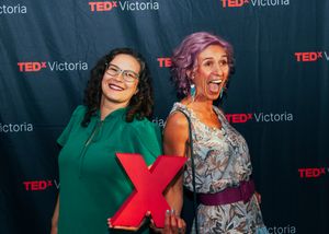 Two women posing and smiling at a TEDx Victoria event backdrop, one holding a large red X prop.