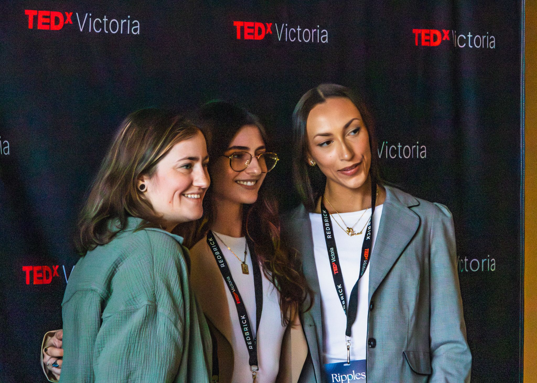 Three women smiling and posing together at a TEDx Victoria event backdrop wearing lanyards.