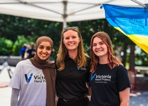 Three women smiling together at a Victoria Hand Project event, with a Ukrainian flag visible in the background.