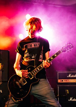 Guitarist performing on stage in a The Nils t-shirt, playing black electric guitar with colorful pink and purple stage lighting.