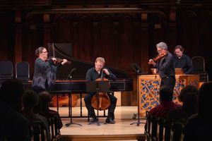 Four classical musicians perform chamber music on stage in an elegant wood-paneled concert hall before a seated audience.