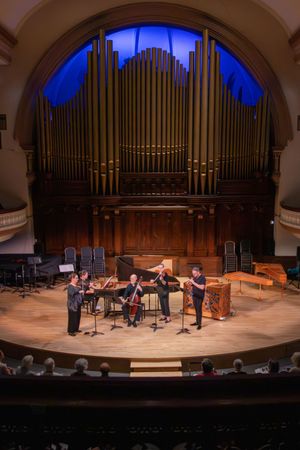 Chamber music ensemble performing on stage before a grand pipe organ in an elegant concert hall.