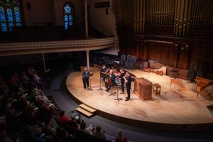 Chamber ensemble performs on a wooden stage in an elegant concert hall with pipe organ and stained glass windows.