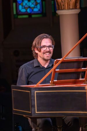 Smiling man with glasses playing a harpsichord in a church with stained glass windows.