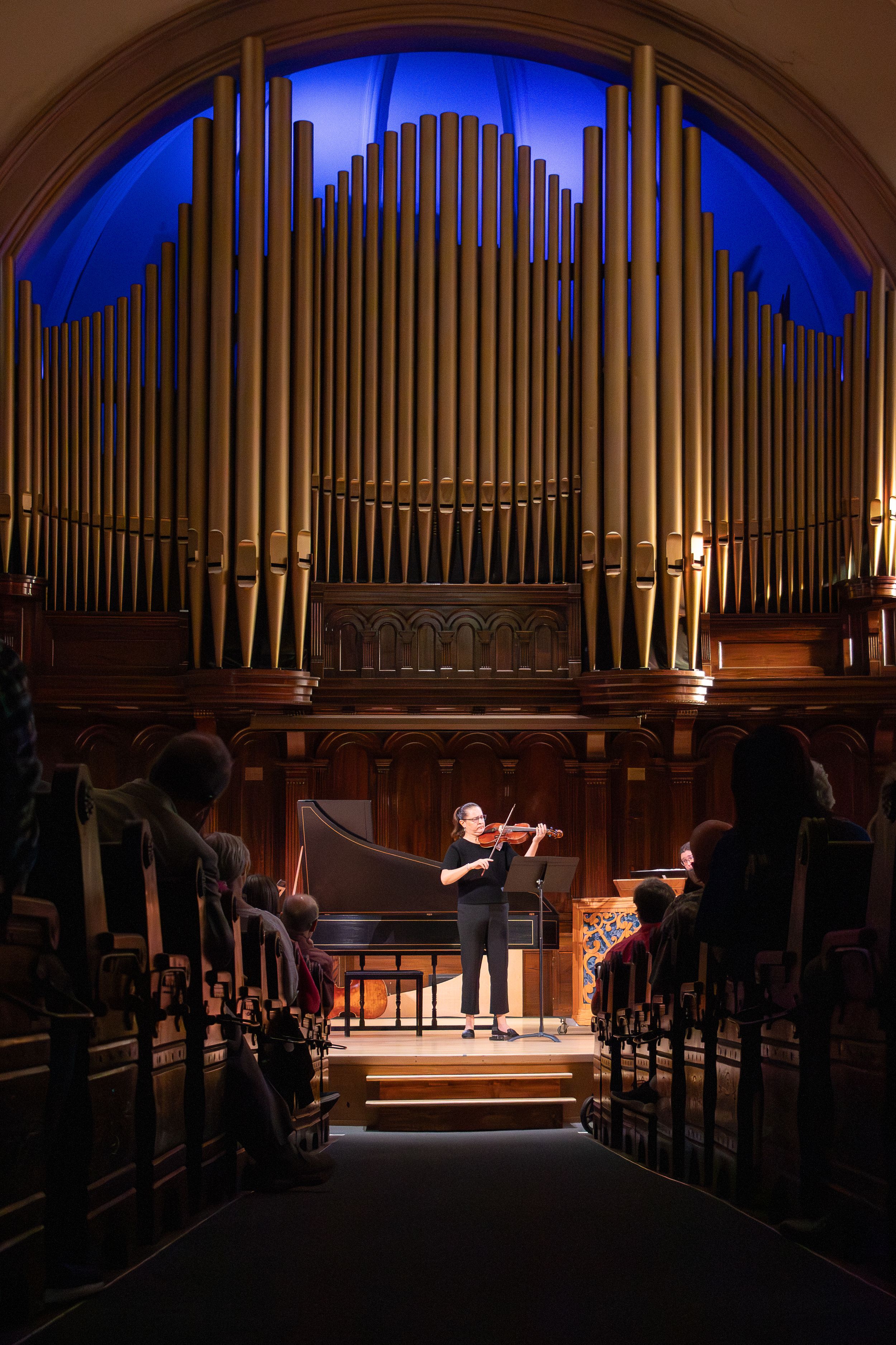 Violinist performs in ornate concert hall with grand pipe organ backdrop, small chamber audience seated in wooden pews.