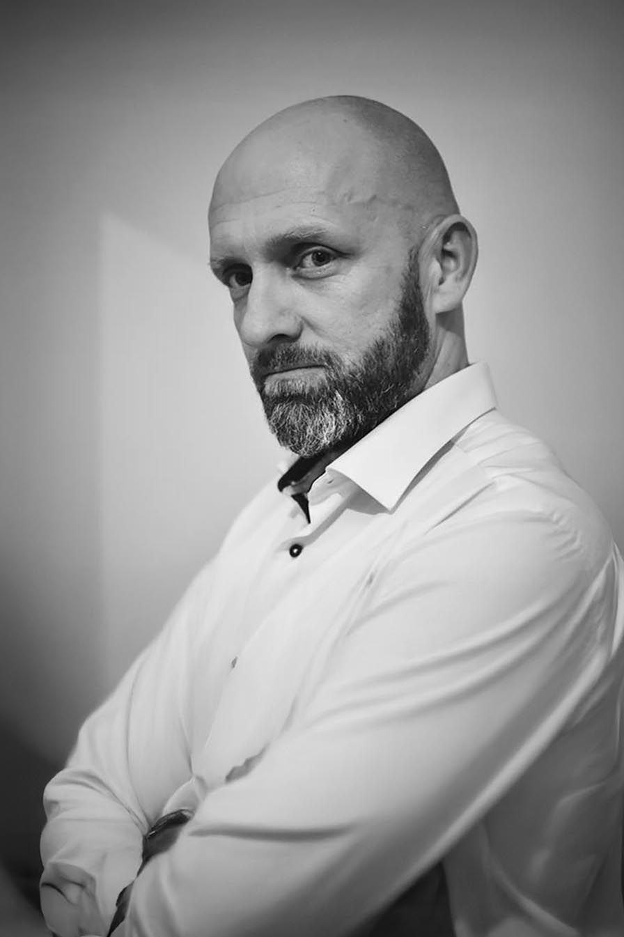 Black and white portrait of a bald man with a beard, arms crossed, wearing a white shirt.