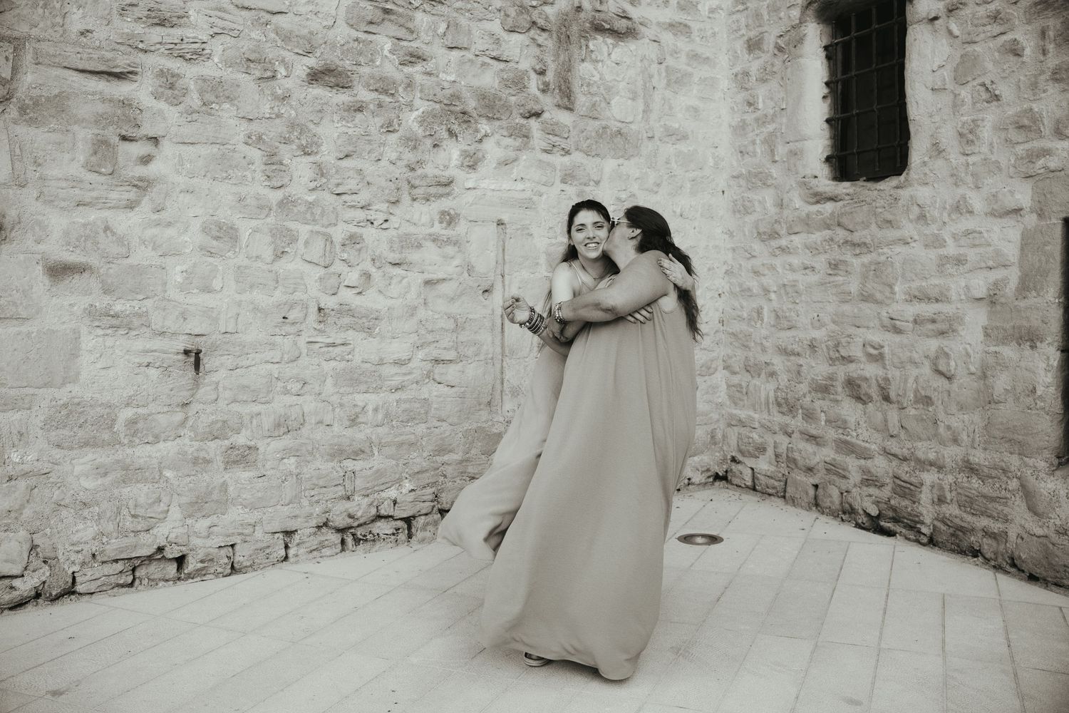 Une mère embrasse sa fille adulte sur la joue, photographiées en pied et en noir et blanc devant les remparts de Trets.