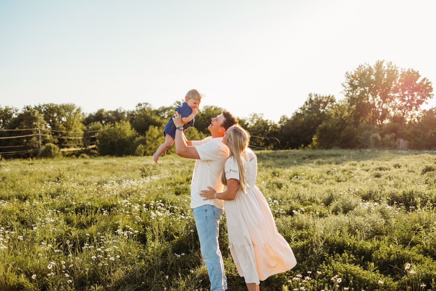 A happy family enjoys a sunny day in a green field, with parents lifting their baby in the air.