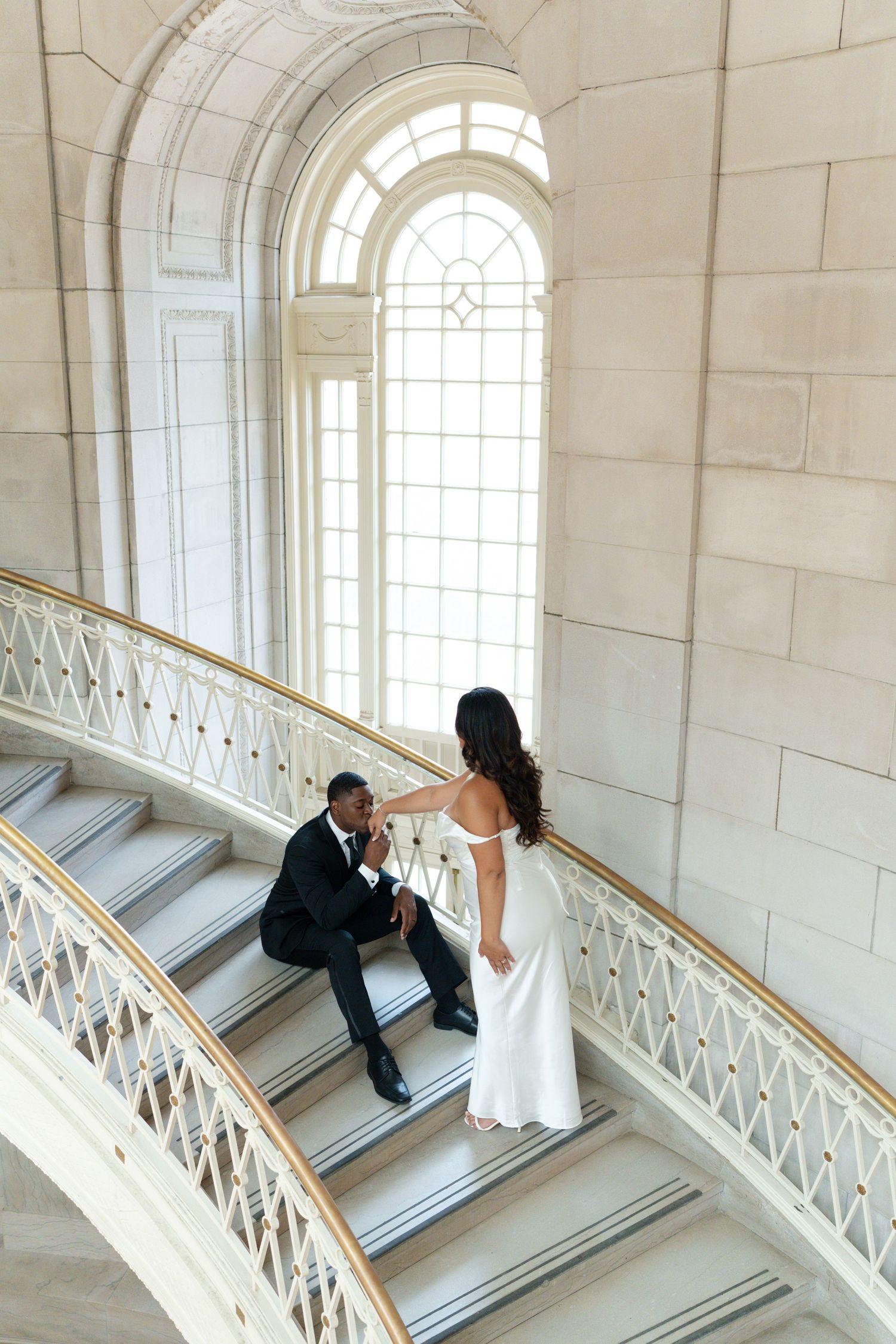 Bride and groom posing on a sweeping white marble staircase beneath a grand arched window.