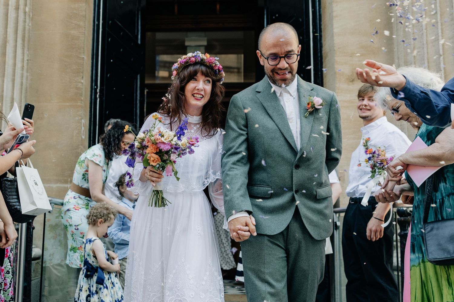 Bride and groom exit church holding hands as guests throw confetti, bride wearing floral crown and white dress.