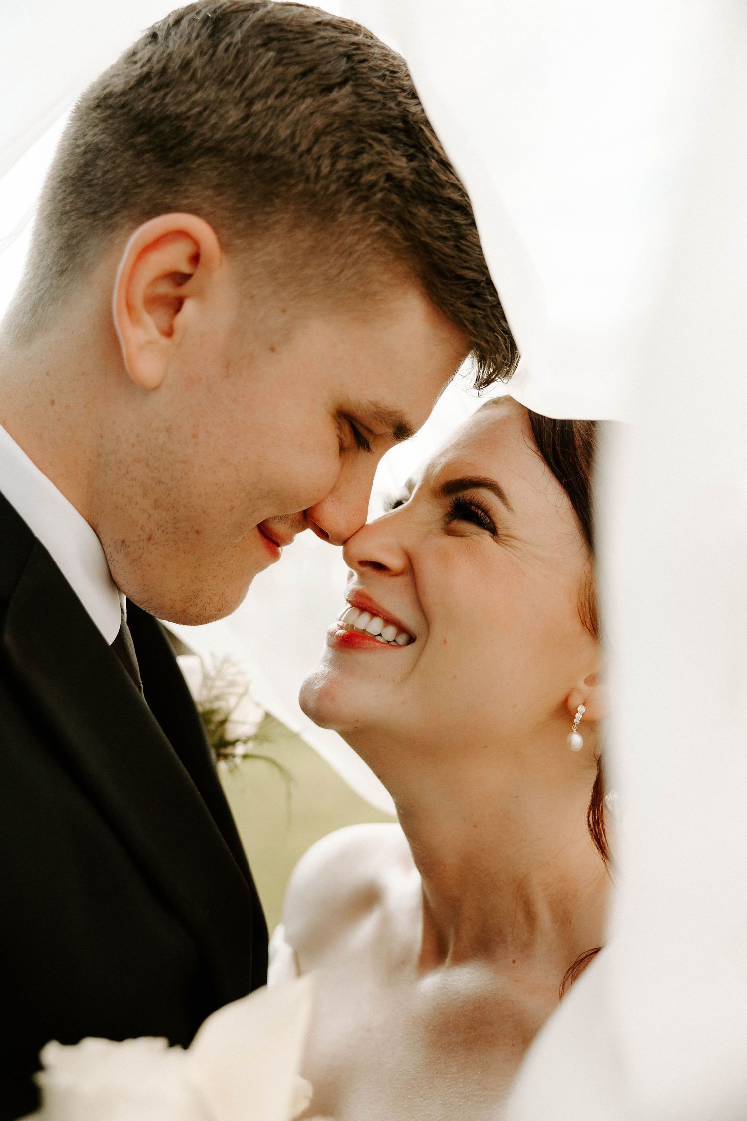 A smiling bride and groom touch foreheads tenderly under a white veil, with a bouquet of white roses in the foreground.