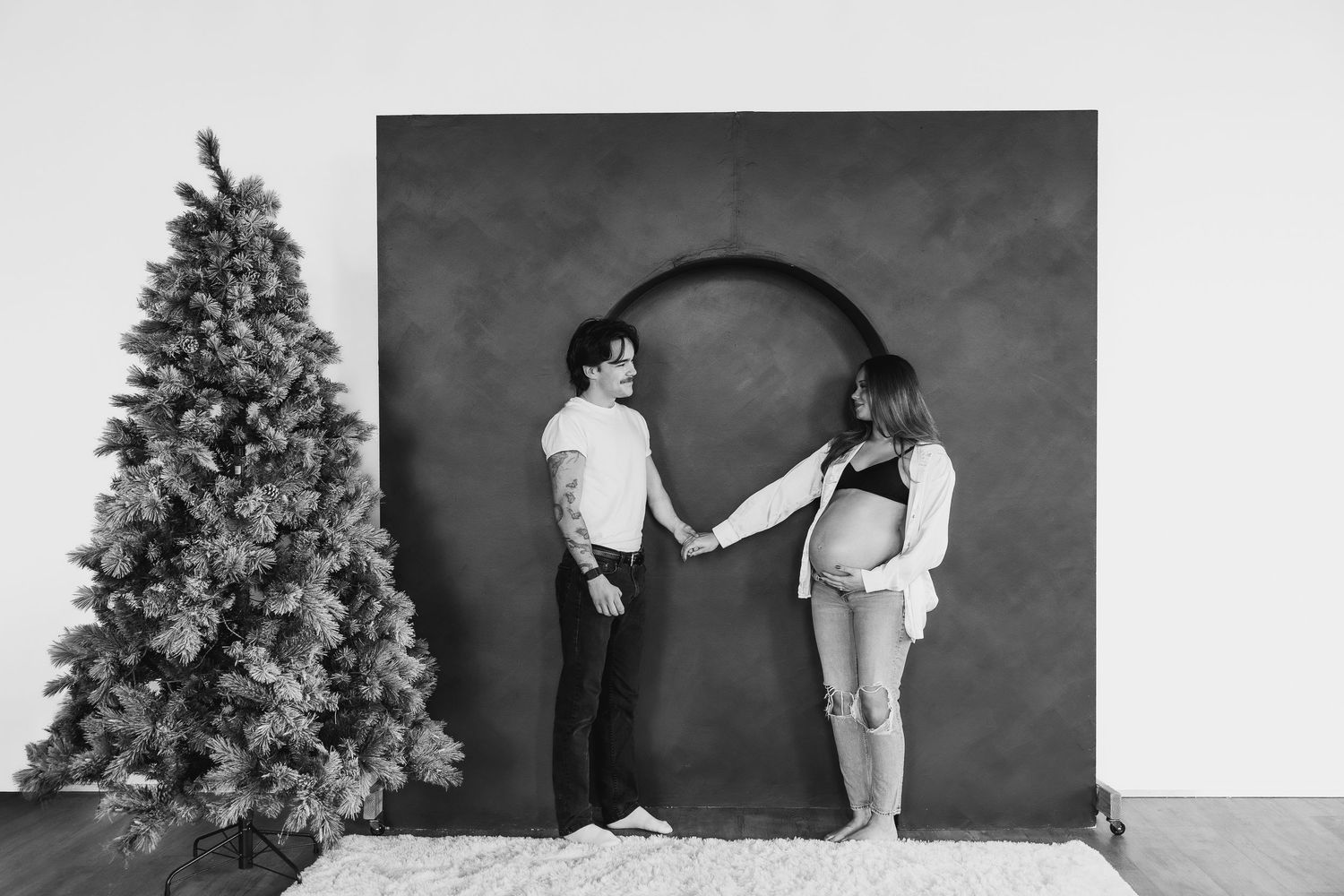 Black and white photo of a couple holding hands near a Christmas tree, with the woman visibly pregnant.