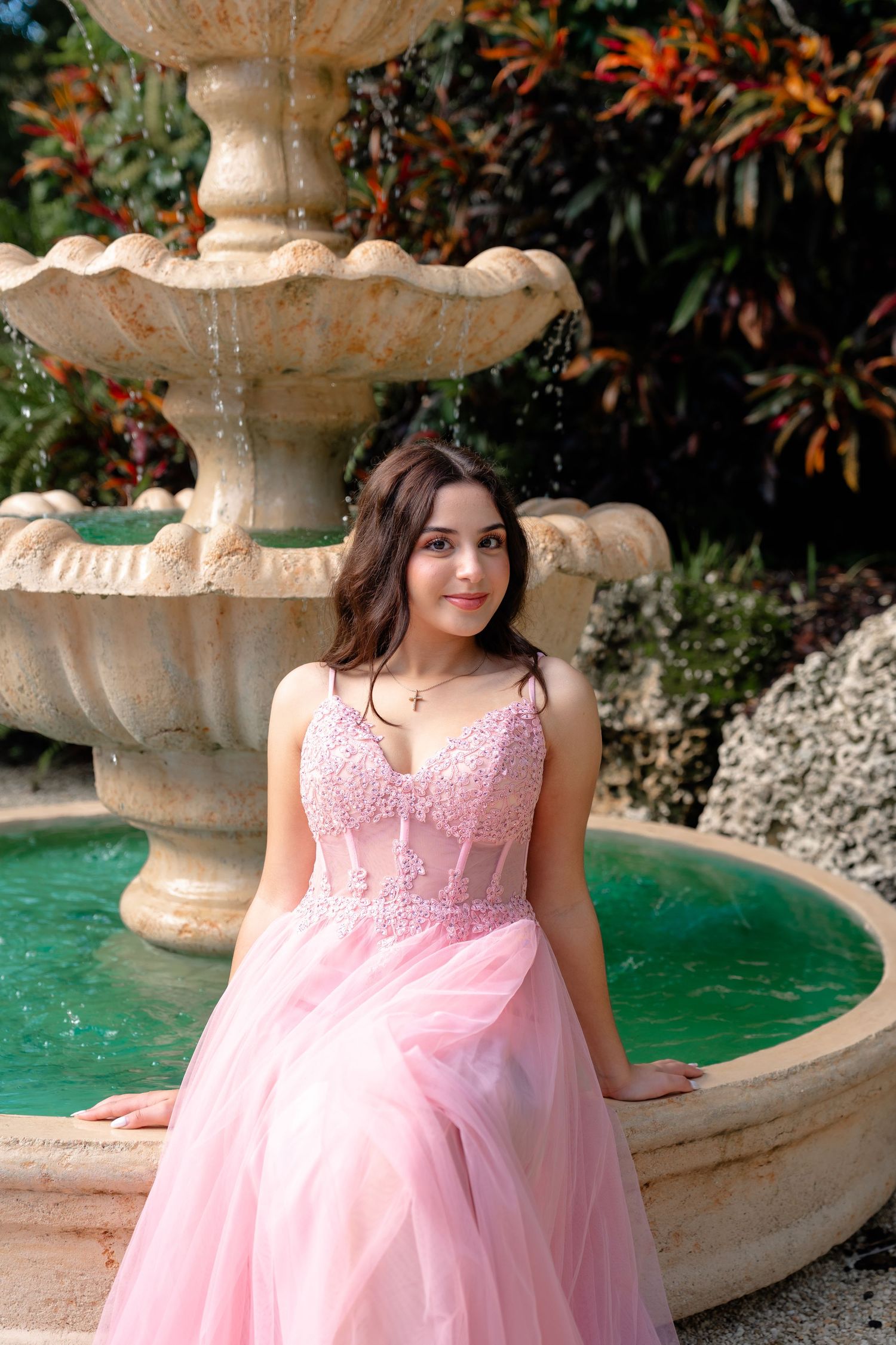 Young woman in pink lace ball gown poses elegantly beside a stone tiered garden fountain outdoors.