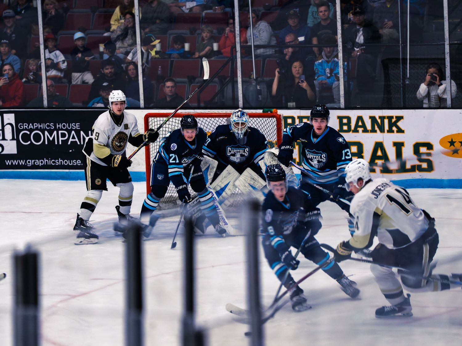 A navy Madison Capitols goaltender and skaters defend the crease as white-and-gold Lumberjacks players crash the net during a scramble.