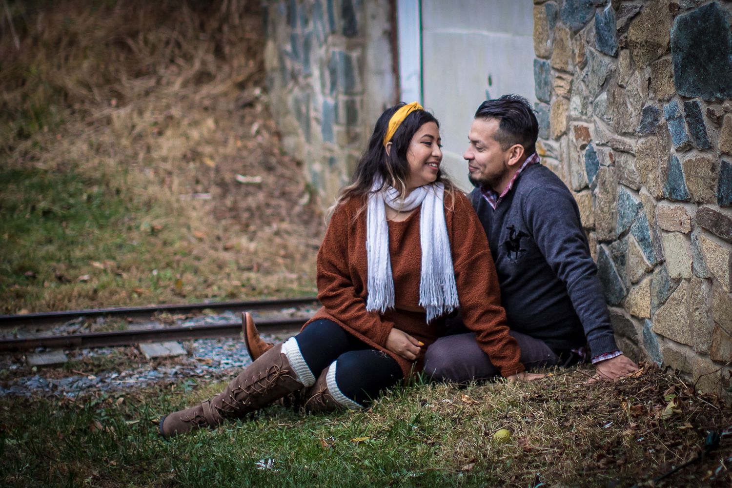 Couple sitting together on grass by stone wall and railroad tracks, sharing a romantic moment outdoors.