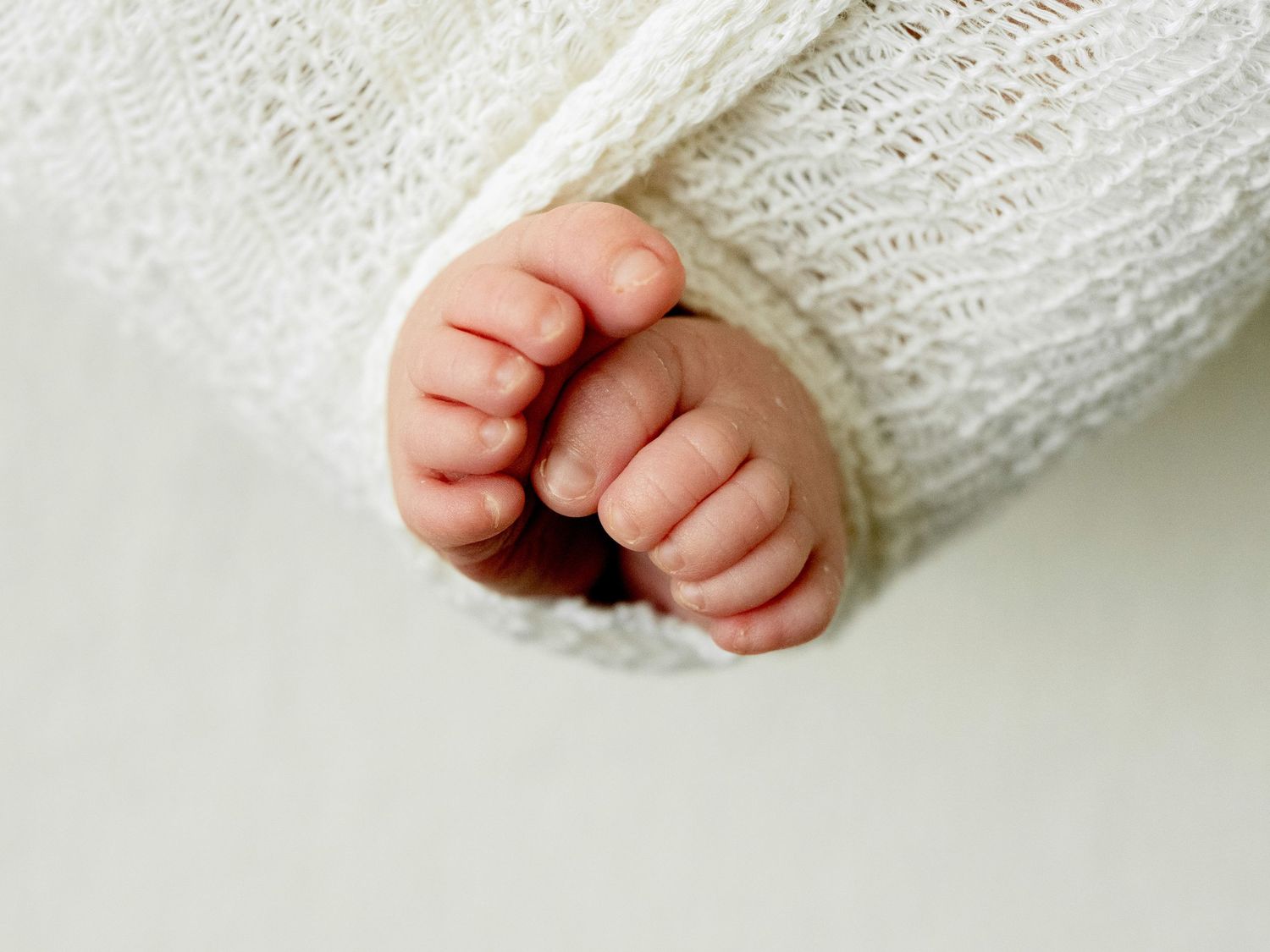 Close-up of tiny newborn baby feet wrapped in a soft white knit blanket on a light background.