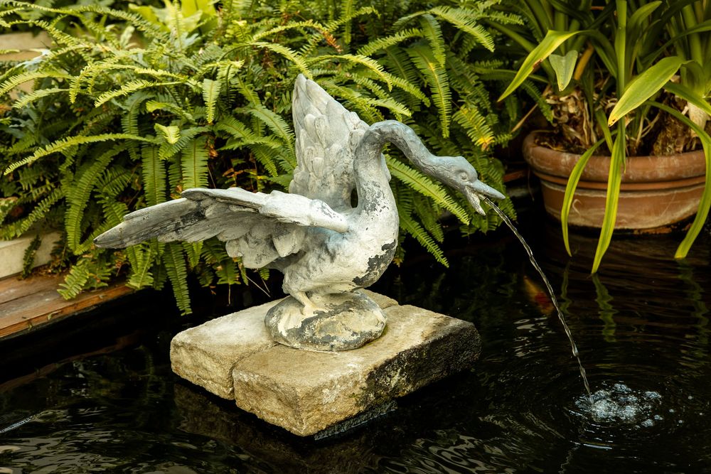 Bronze heron statue with spread wings on stone base beside a garden pond fountain surrounded by lush ferns.