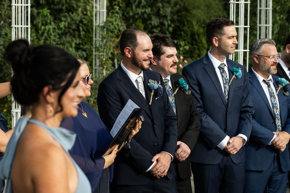 Groomsmen in navy suits stand at outdoor wedding ceremony with lush greenery backdrop.