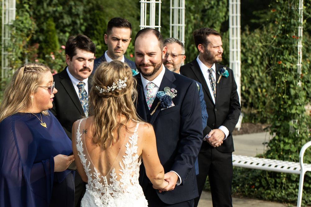 Groom smiling at bride during outdoor wedding ceremony with groomsmen standing in background.