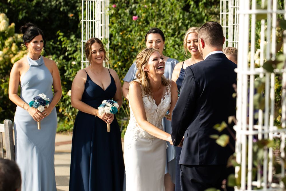Bride laughing joyfully with bridesmaids in blue dresses during outdoor wedding ceremony surrounded by lush greenery.