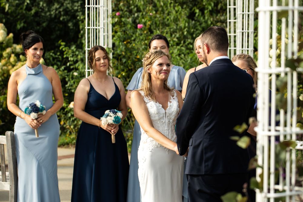 Bride smiling at groom during outdoor wedding ceremony with bridesmaids in blue dresses holding bouquets.