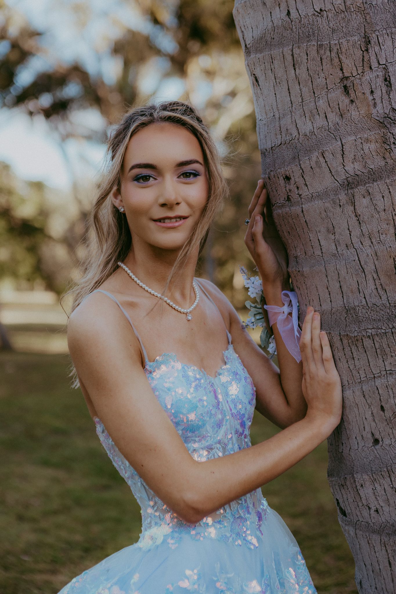 Blonde teen in blue floral formal dress leaning against palm tree at golden hour.