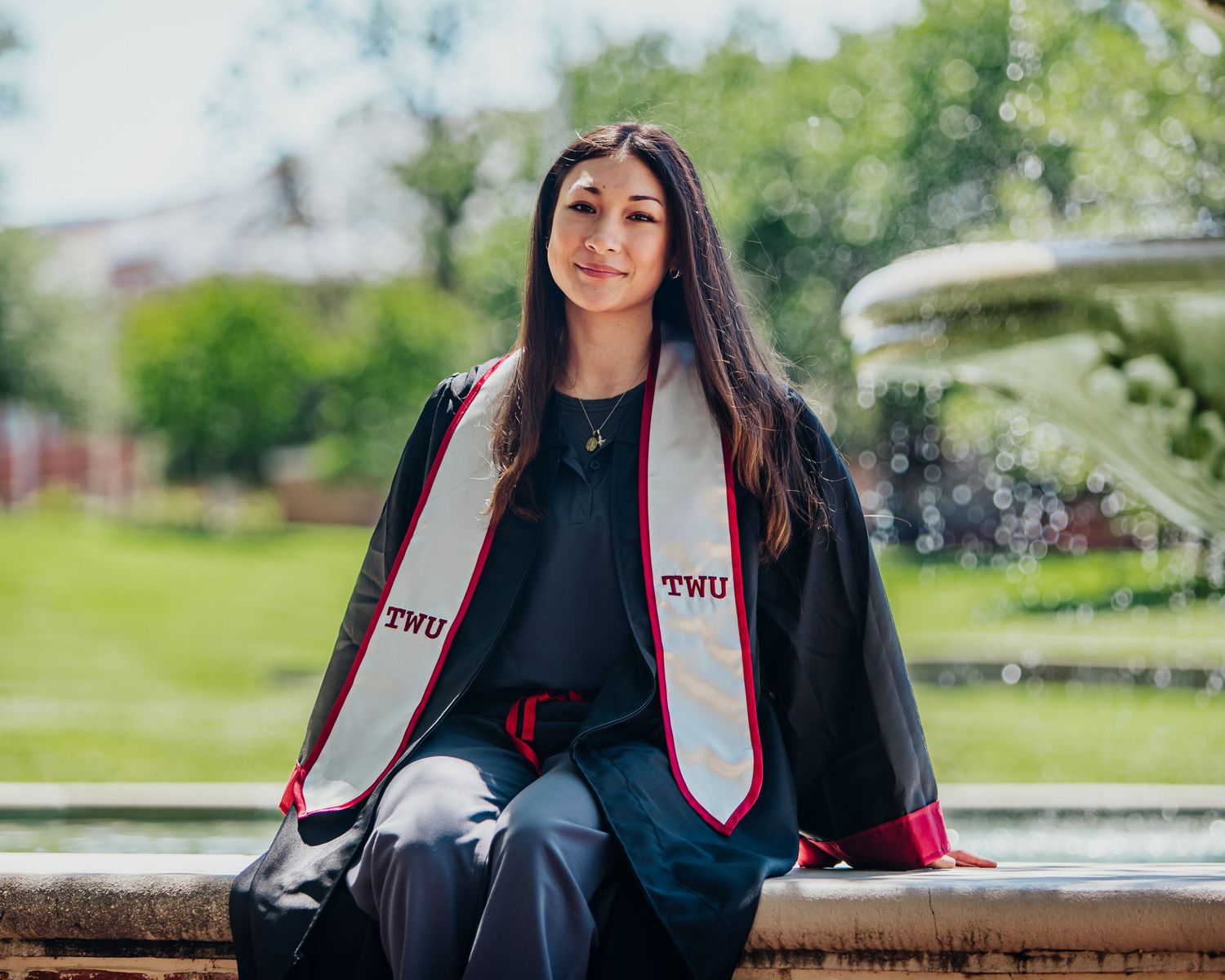 College graduate in cap and gown posing outdoors by a campus fountain