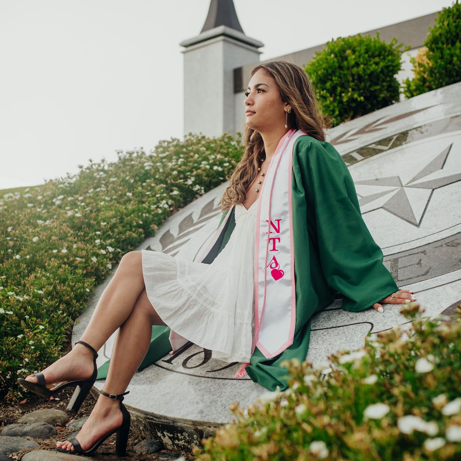 College graduate in a green graduation gown posing among flowers on campus