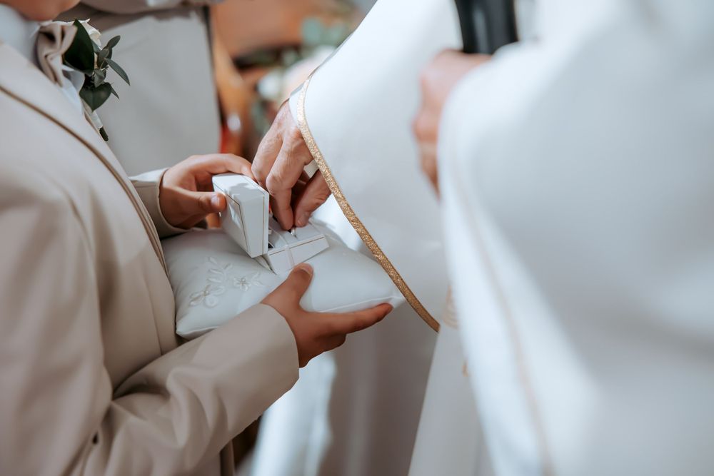 Priest blessing wedding rings on a white pillow during a church ceremony.