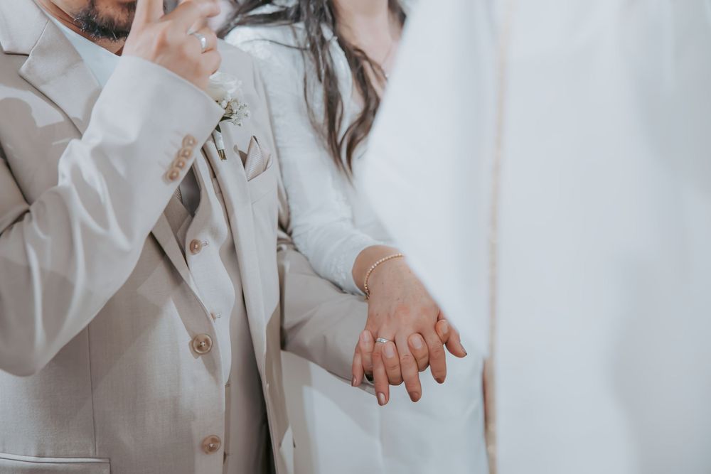 Close-up of newlyweds holding hands, bride in white gown and groom in beige suit with boutonniere.