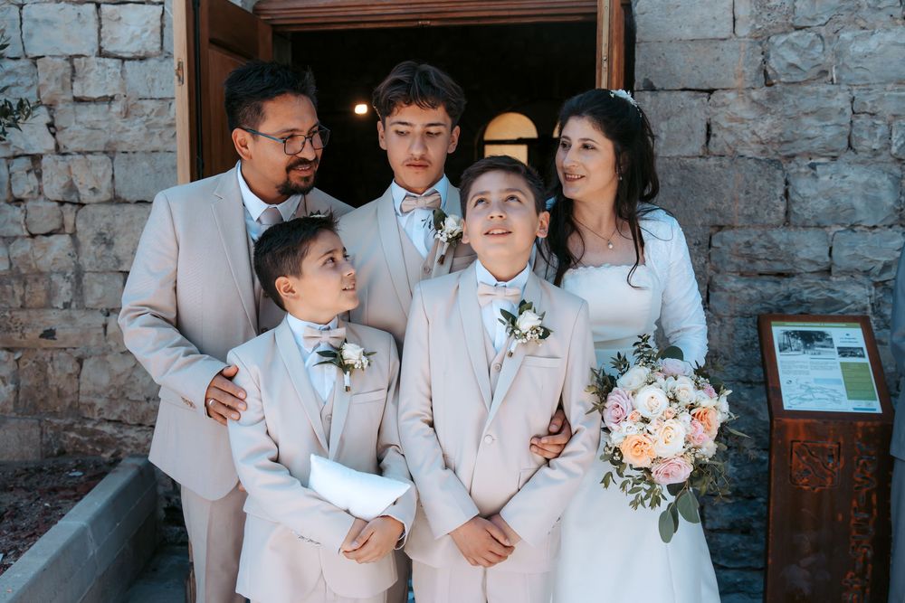 Happy wedding family portrait with groom, bride holding bouquet, and three boys in matching beige suits.