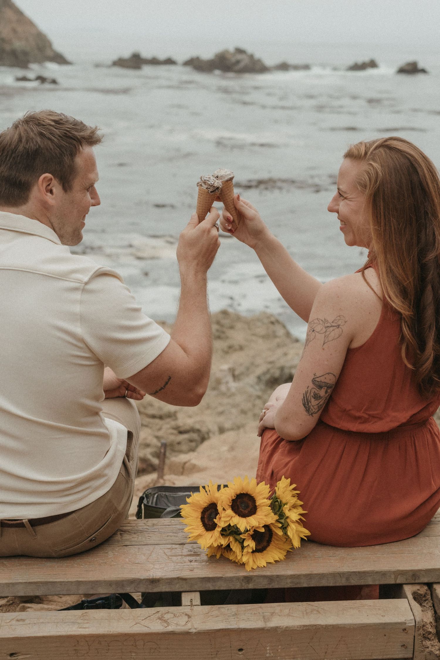 Couple sitting on wooden pallet at rocky beach, toasting with shells, sunflowers nearby, ocean backdrop.