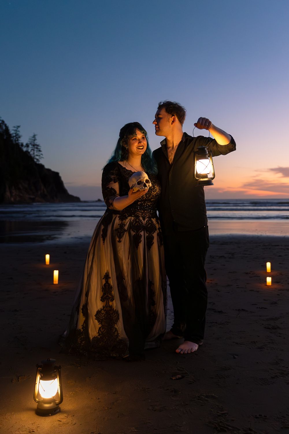 Couple holding lanterns on a beach at sunset, surrounded by candles, woman in elegant black gown, romantic evening setting.