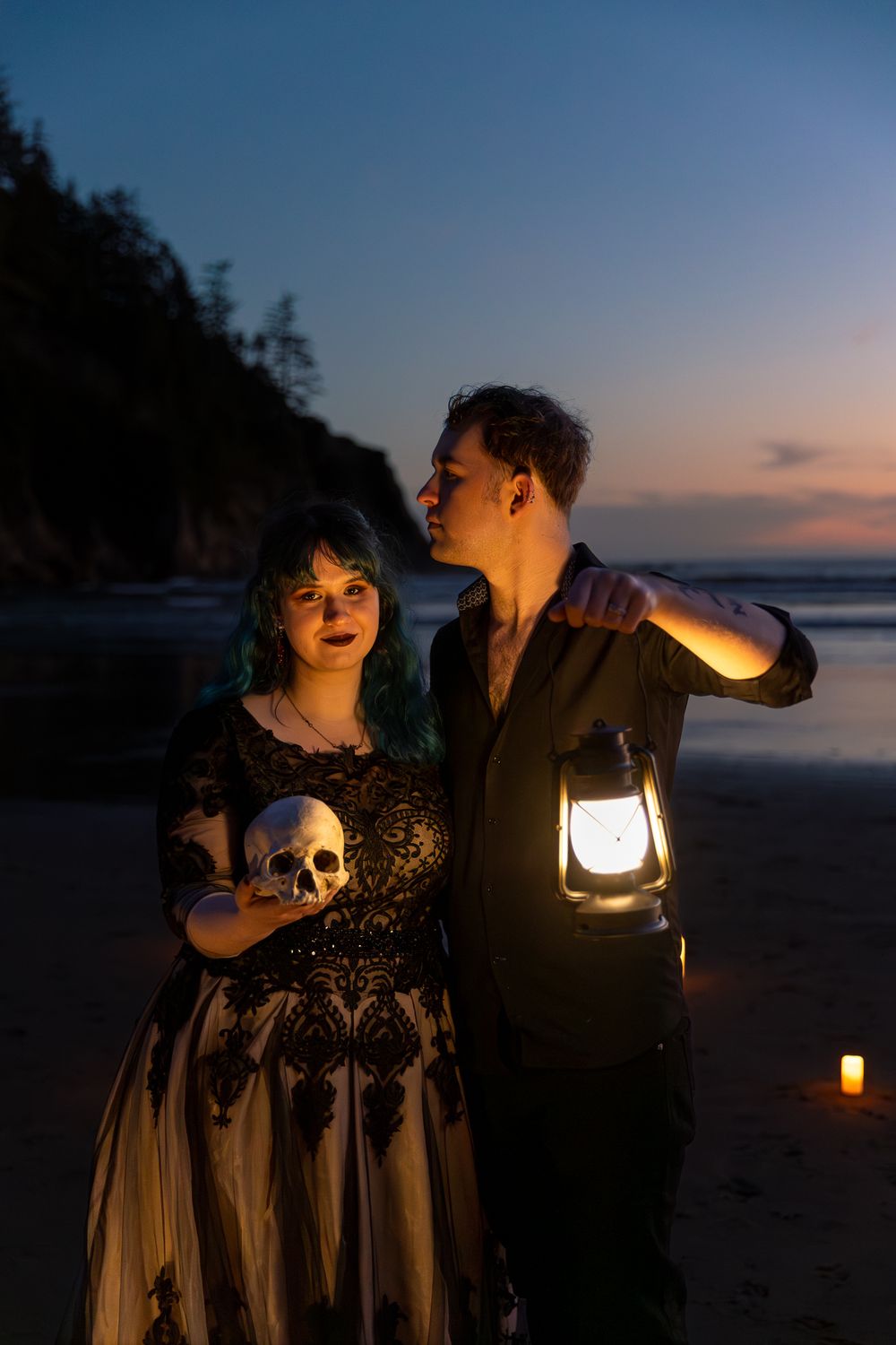 Gothic couple on beach at dusk, woman holding skull in lace dress, man with glowing lantern, candlelit romantic scene.