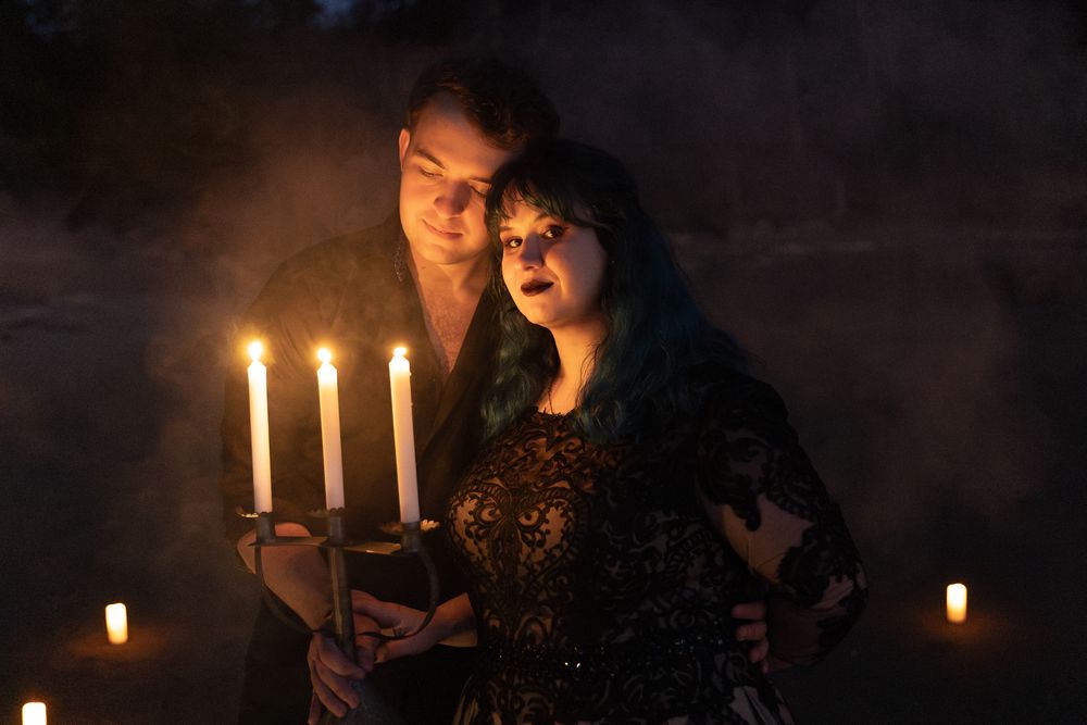 Gothic couple holding candelabra with lit candles in misty dark atmosphere, woman with teal hair in lace dress.