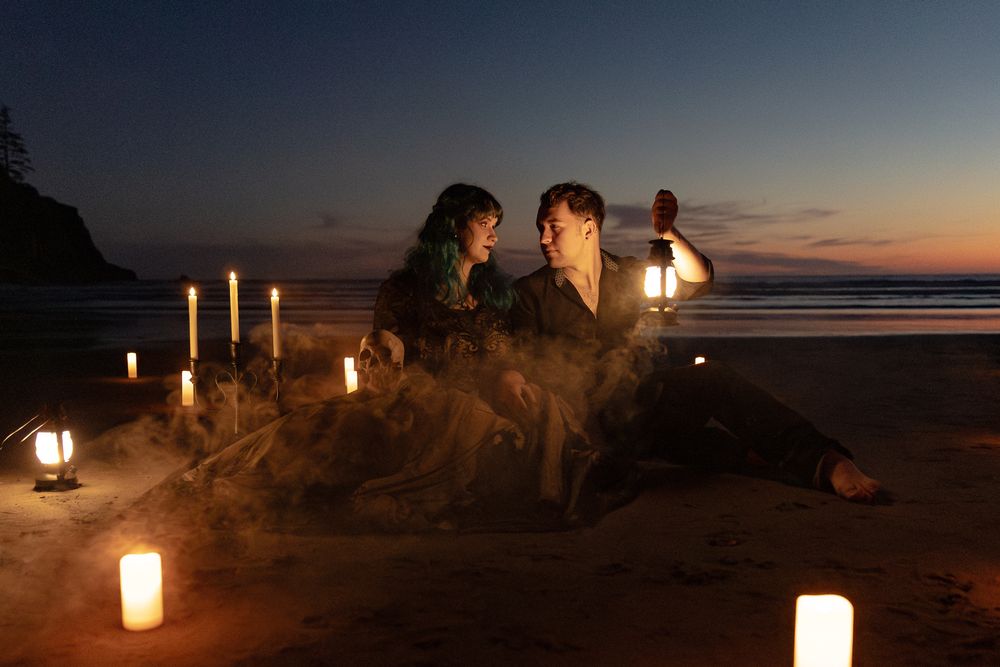 Two people sit surrounded by glowing candles on a beach at dusk, one holding a lantern in a romantic setting.