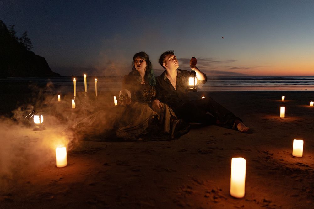 Two people sit on a candlelit beach at dusk surrounded by glowing candles and mystic smoke in a romantic twilight setting.
