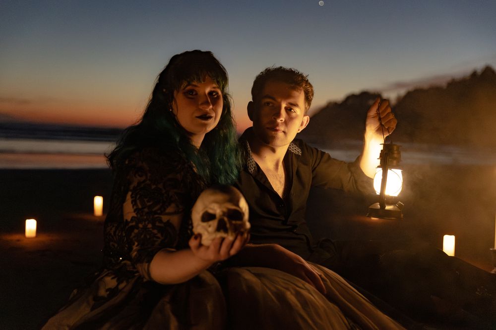 Two people pose at dusk with candles, a skull, and a lantern in a dramatic Halloween-themed outdoor scene.