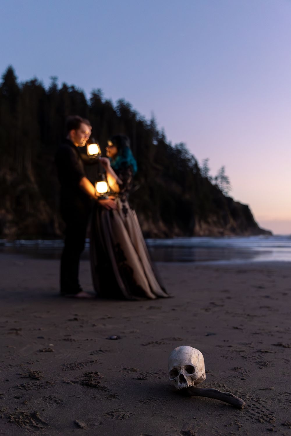 Couple holding lanterns on a dark beach at dusk with a skull in the foreground and forested cliffs behind.