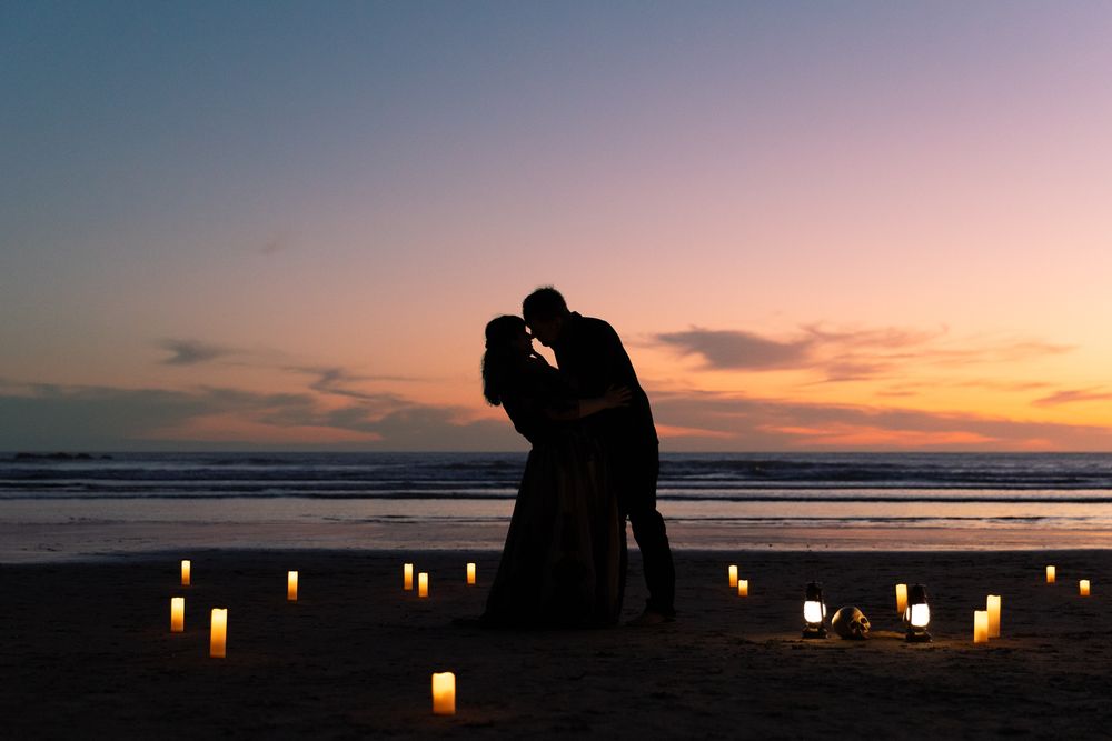 Romantic couple silhouette kissing on beach at sunset surrounded by glowing candles on sand.