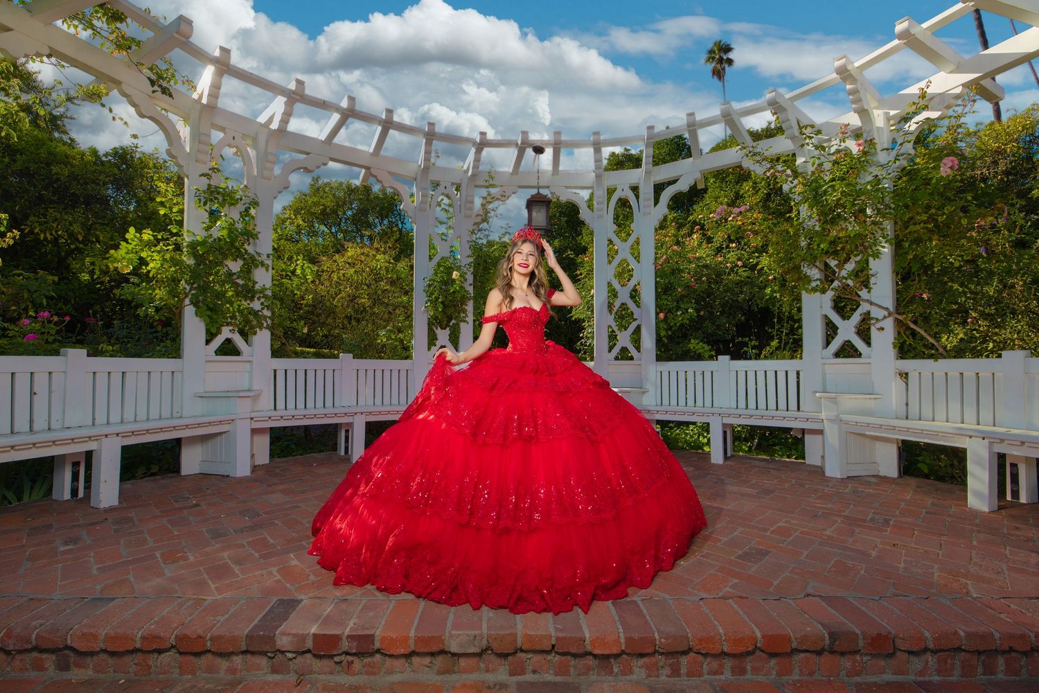 Young woman in a stunning red ball gown poses under a white garden gazebo with lush greenery and blue sky.