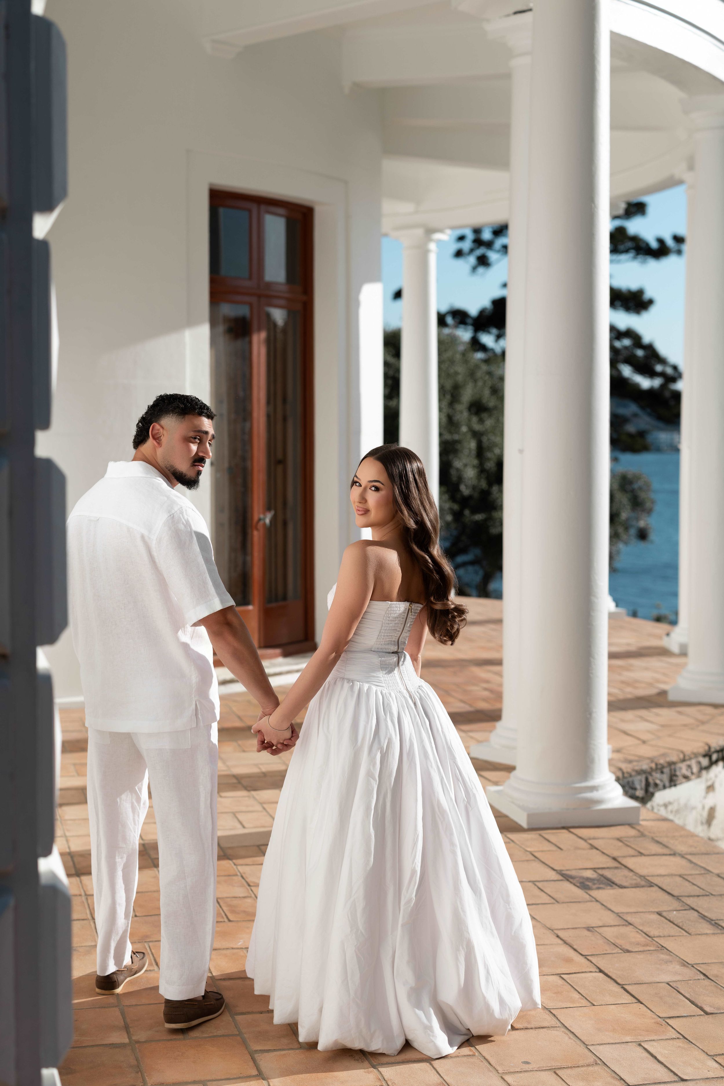 Couple in white wedding attire holding hands on elegant columned terrace overlooking the ocean.