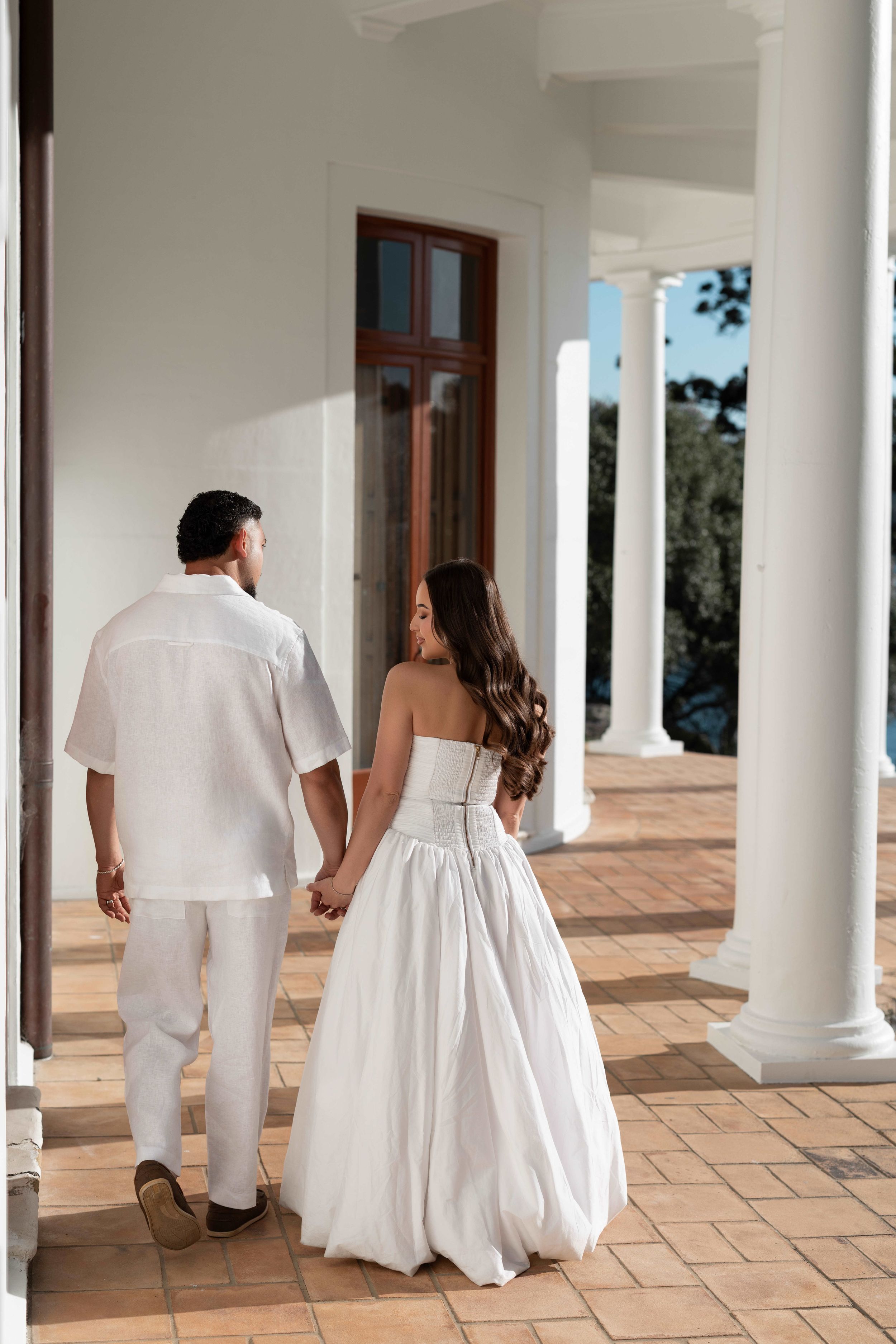 A couple in white attire holds hands while walking along a sunlit colonial-style veranda with white columns.