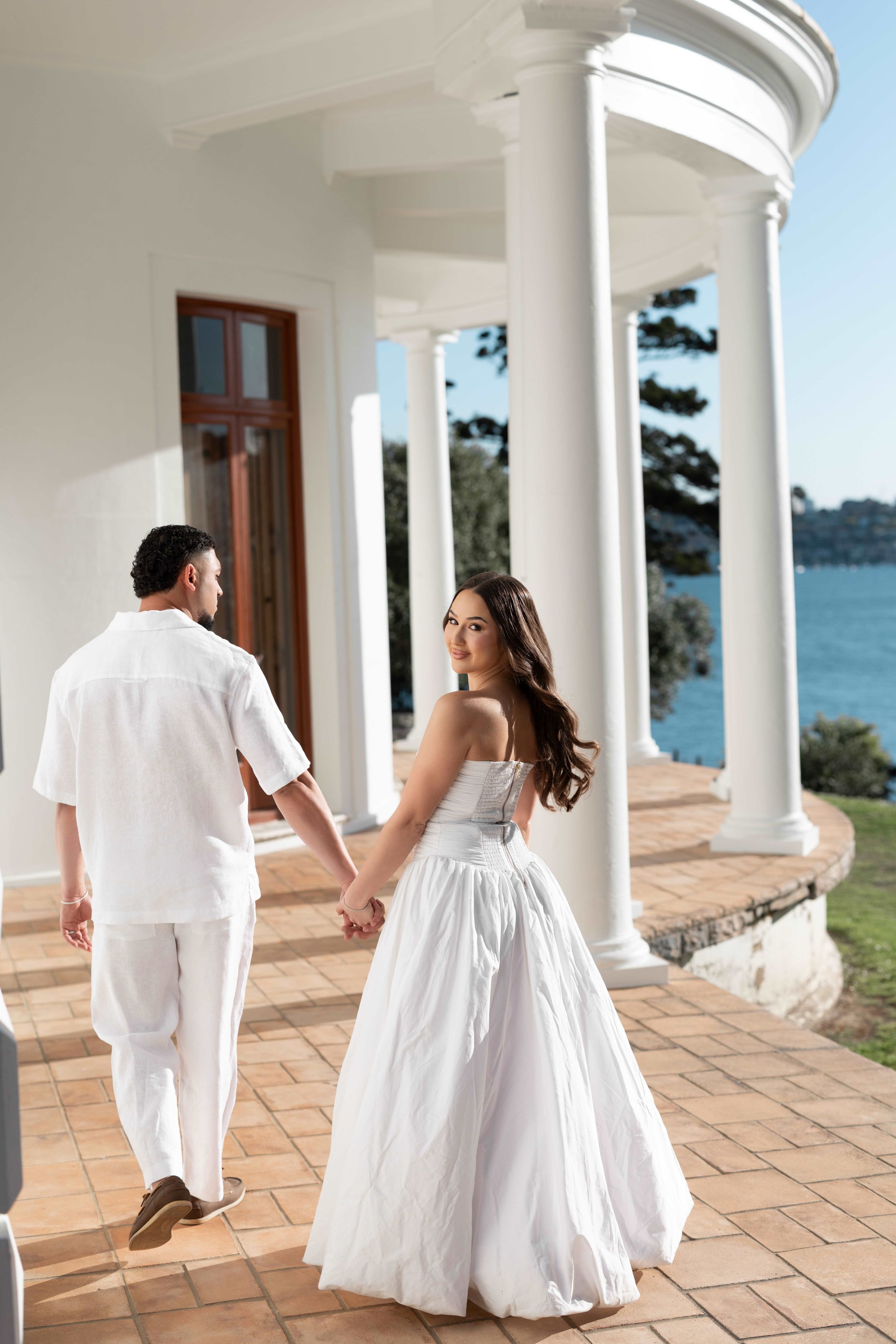 Couple in white attire holding hands on elegant columned veranda overlooking water, bride glancing back.