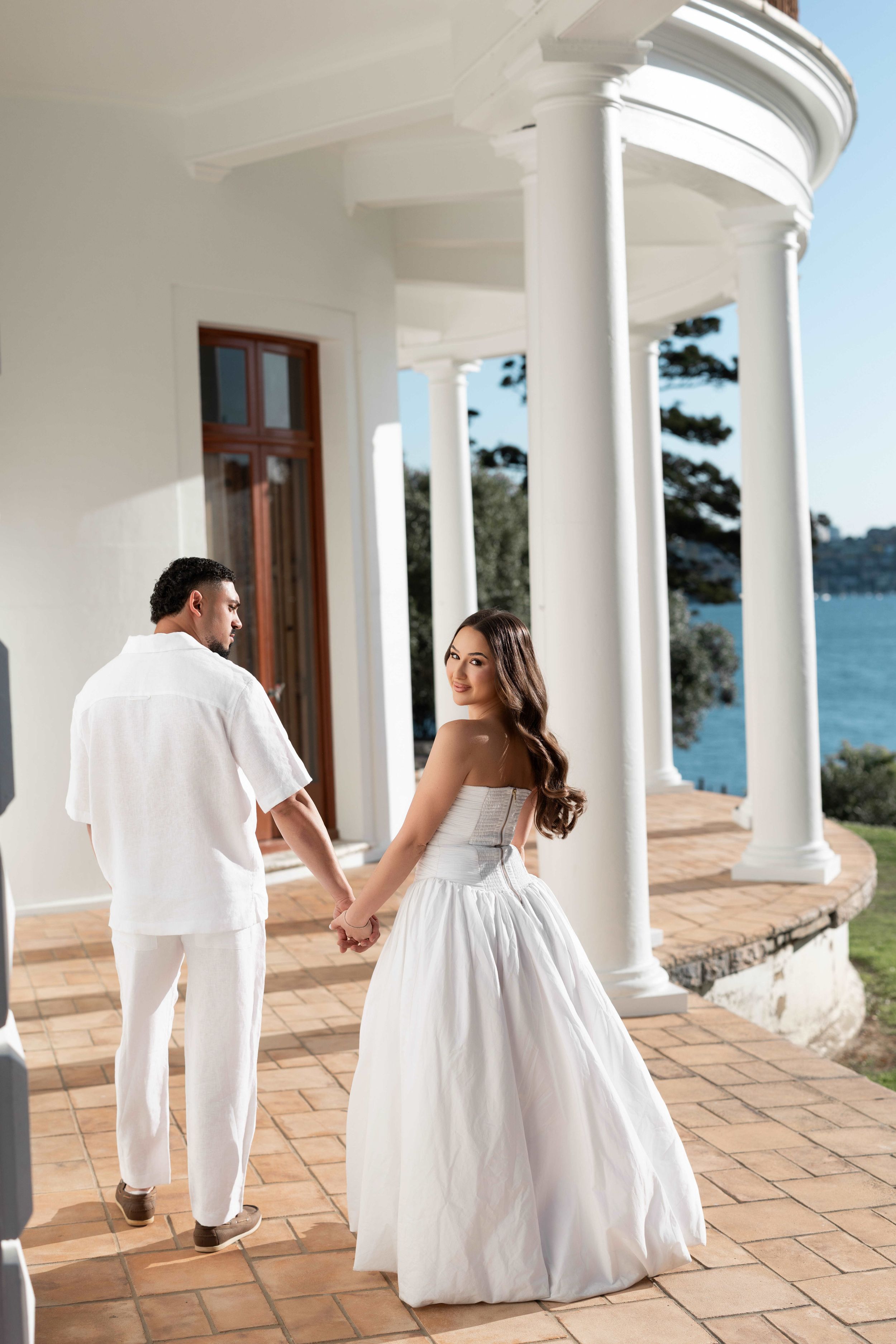 Couple in white attire holding hands on elegant columned terrace overlooking water at a wedding venue.