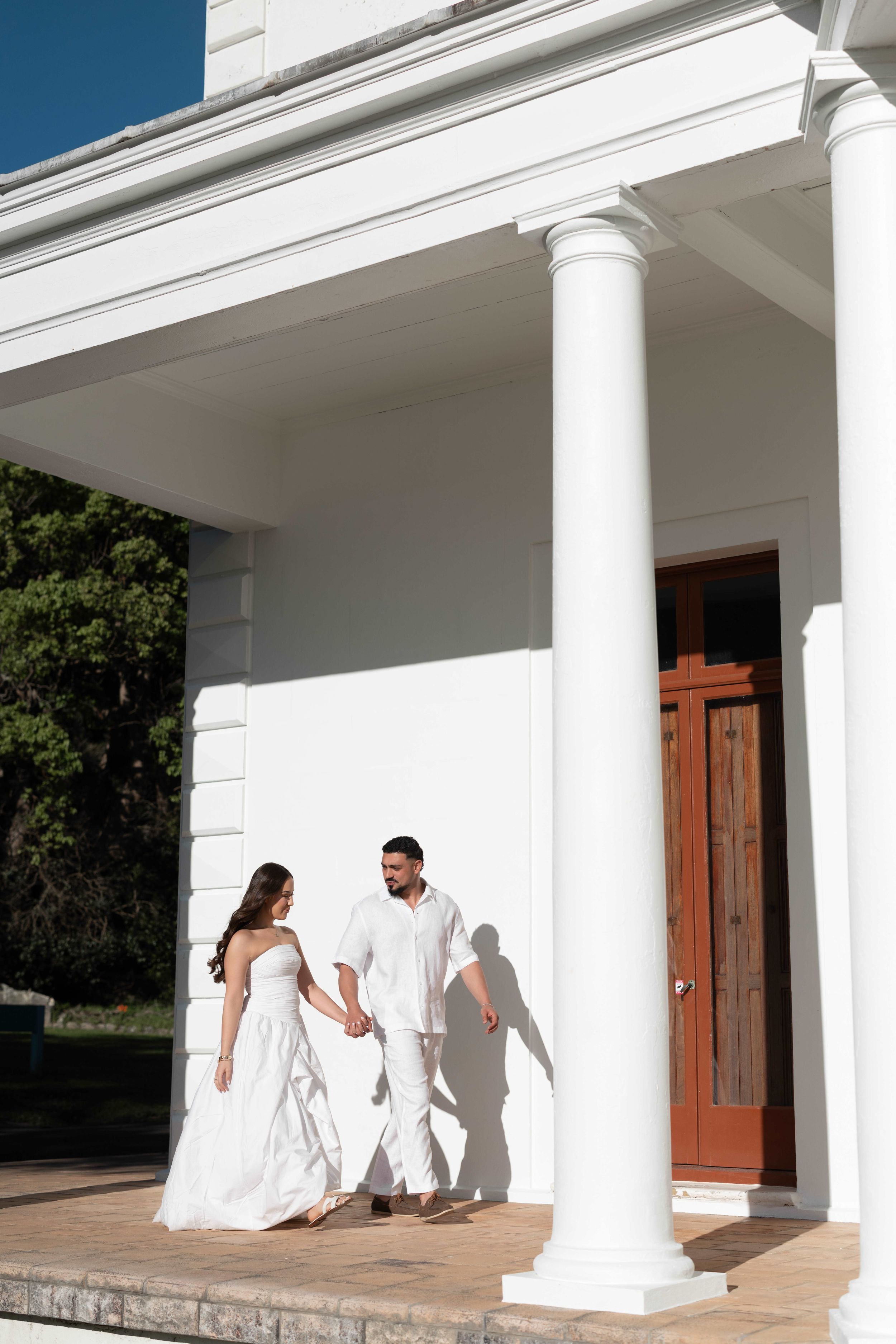 Couple in white outfits holding hands on the porch of an elegant white colonial building with tall columns.