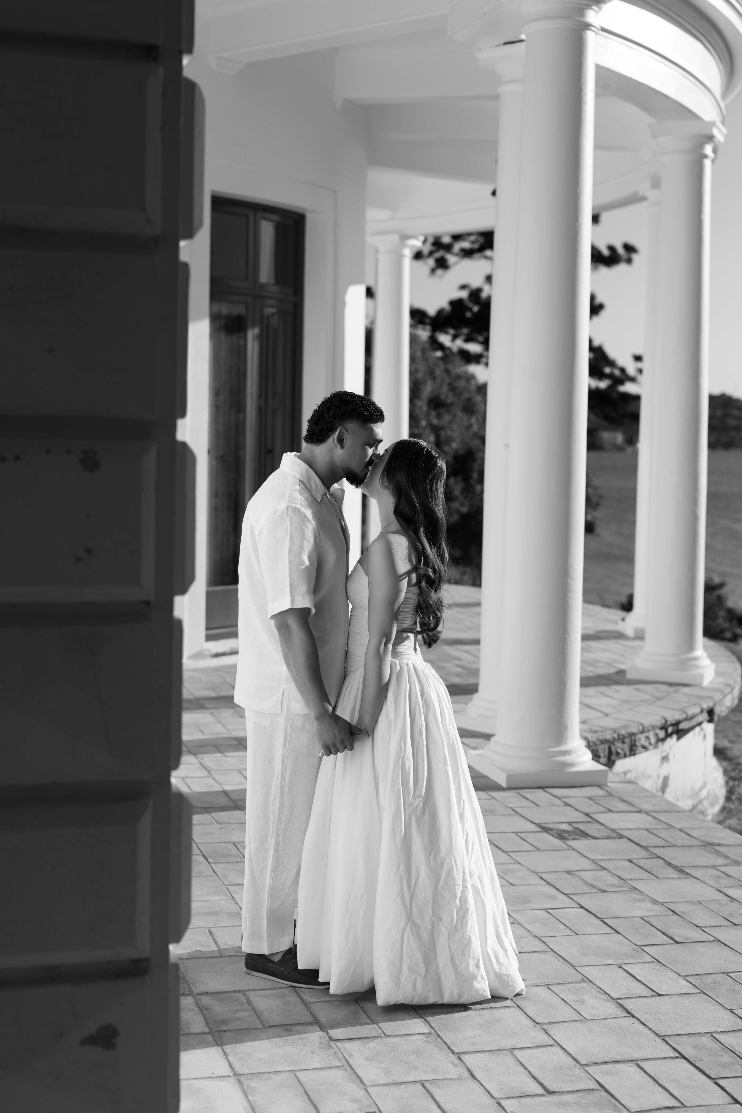 Black and white photo of a couple sharing a romantic moment on a elegant columned porch in wedding attire.