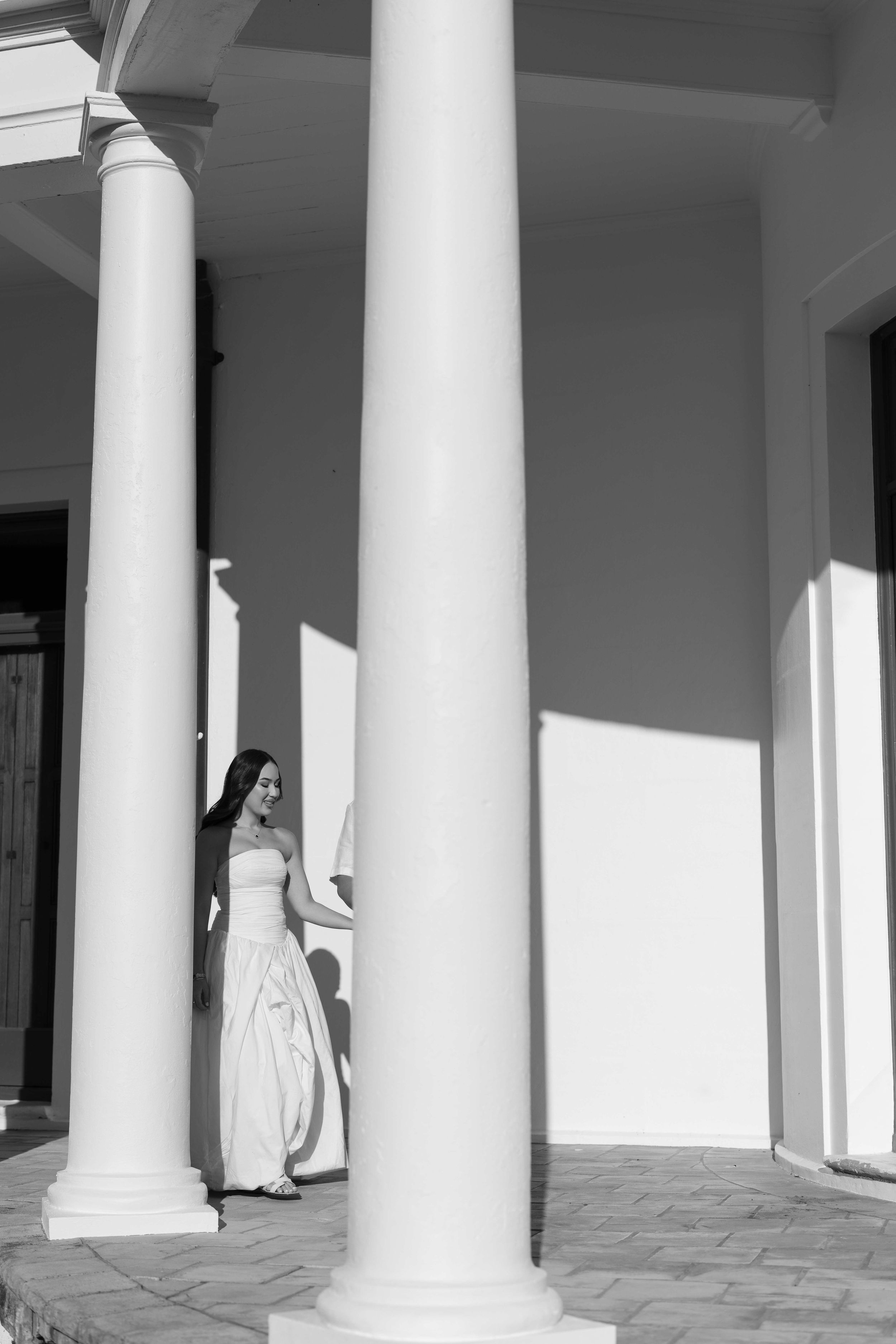 Black and white photo of a bride in a white gown standing between large white columns on a sunlit porch.