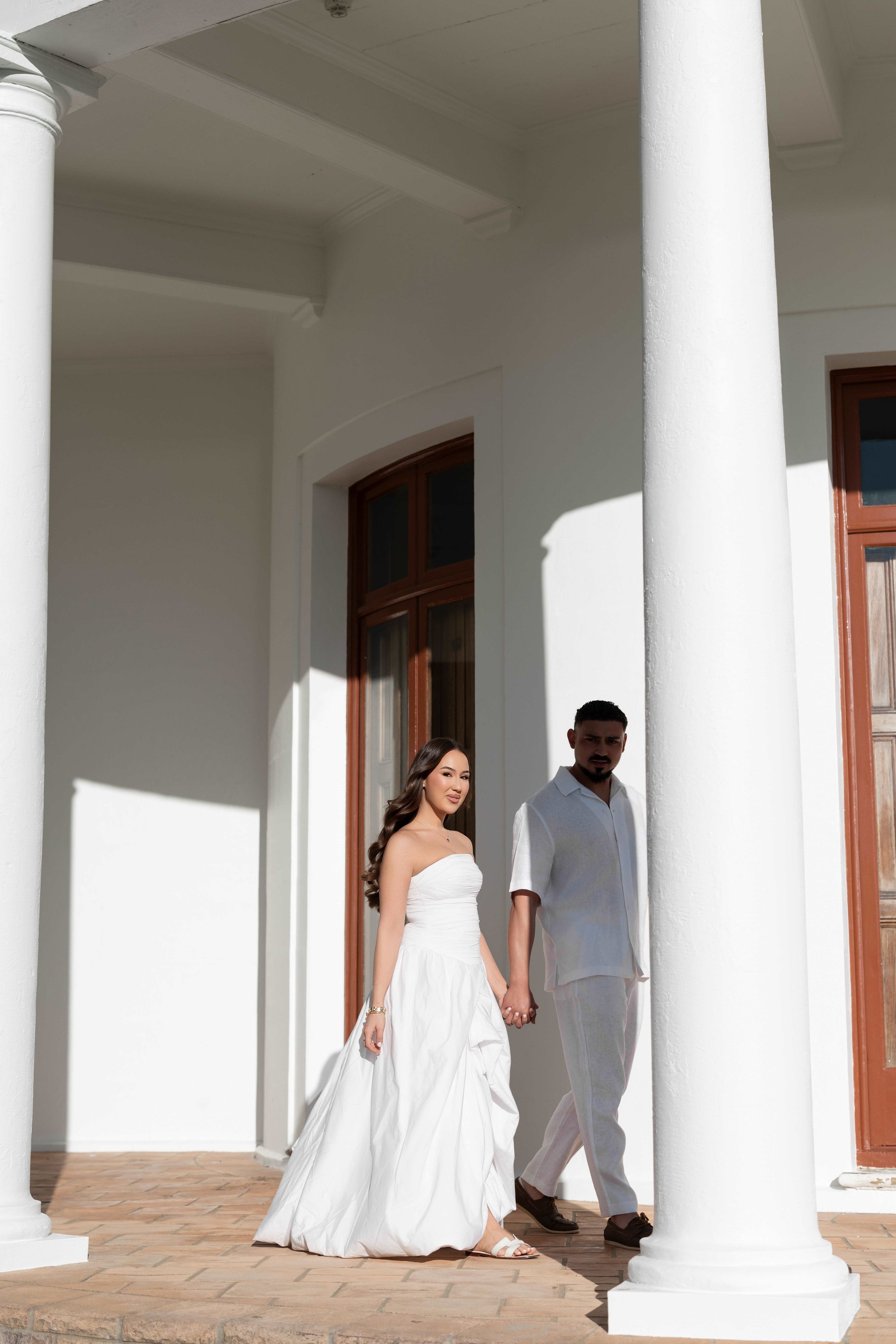 Interracial couple holding hands on a sunlit colonial building porch, bride in white strapless gown, groom in casual linen attire.